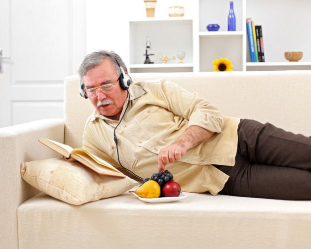 Sénior allongé sur un canapé en train d'écouter de la musique et de manger des fruits