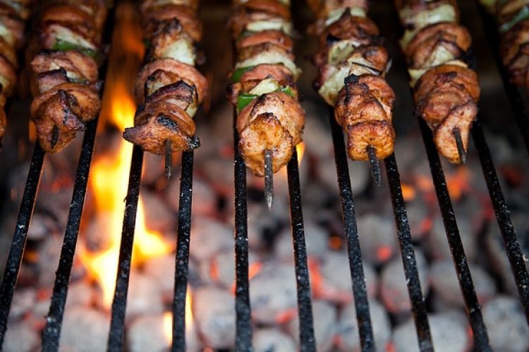 Alignement de cinq brochettes de viandes en train de cuire sur une grille de barbecue