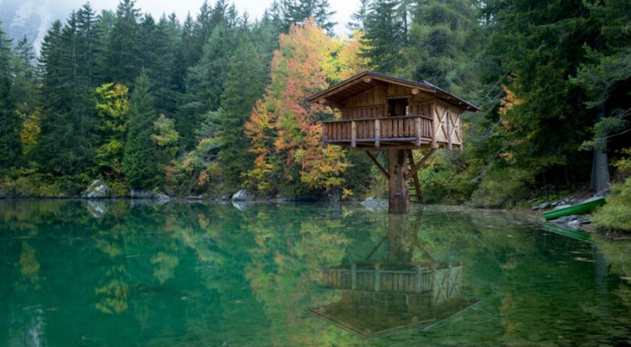 Une maison surplombe un lac, perchée dans les arbres. Son image se reflète sur l'eau.